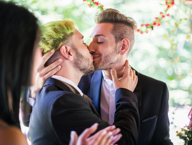 Two gay men passionately kissing in front of a cheering crowd at their Long Island wedding ceremony, officiated by an experienced and inclusive officiant.
