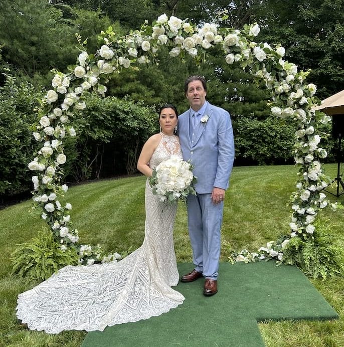 A bride in a white gown and a groom in a light blue suit stand under a circular floral arch outdoors, holding a bouquet of white flowers.