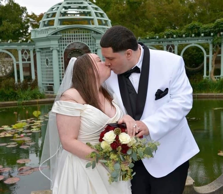 A couple dressed in wedding attire share a kiss in front of a pond. The bride holds a bouquet of red and white flowers, and the groom wears a white tuxedo jacket.