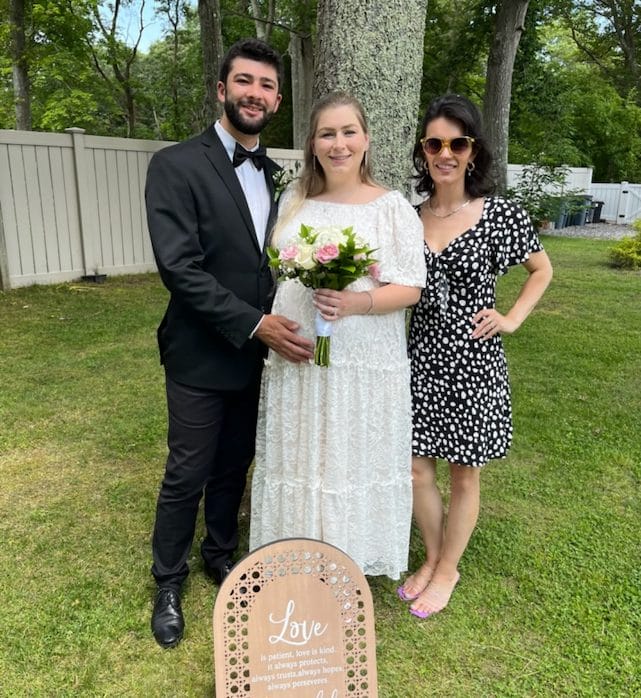 A couple in wedding attire stands next to a woman in a black dress with white patterns. The bride holds a bouquet. A wooden sign with wedding text is in front of them. They are outdoors with trees and a fence in the background.